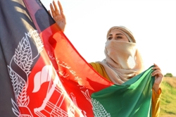 [ai] A woman wearing a light-colored scarf holds the Afghan flag, which features the national emblem, in a field during sunset. She is waving the flag, showcasing a sense of pride in her heritage.